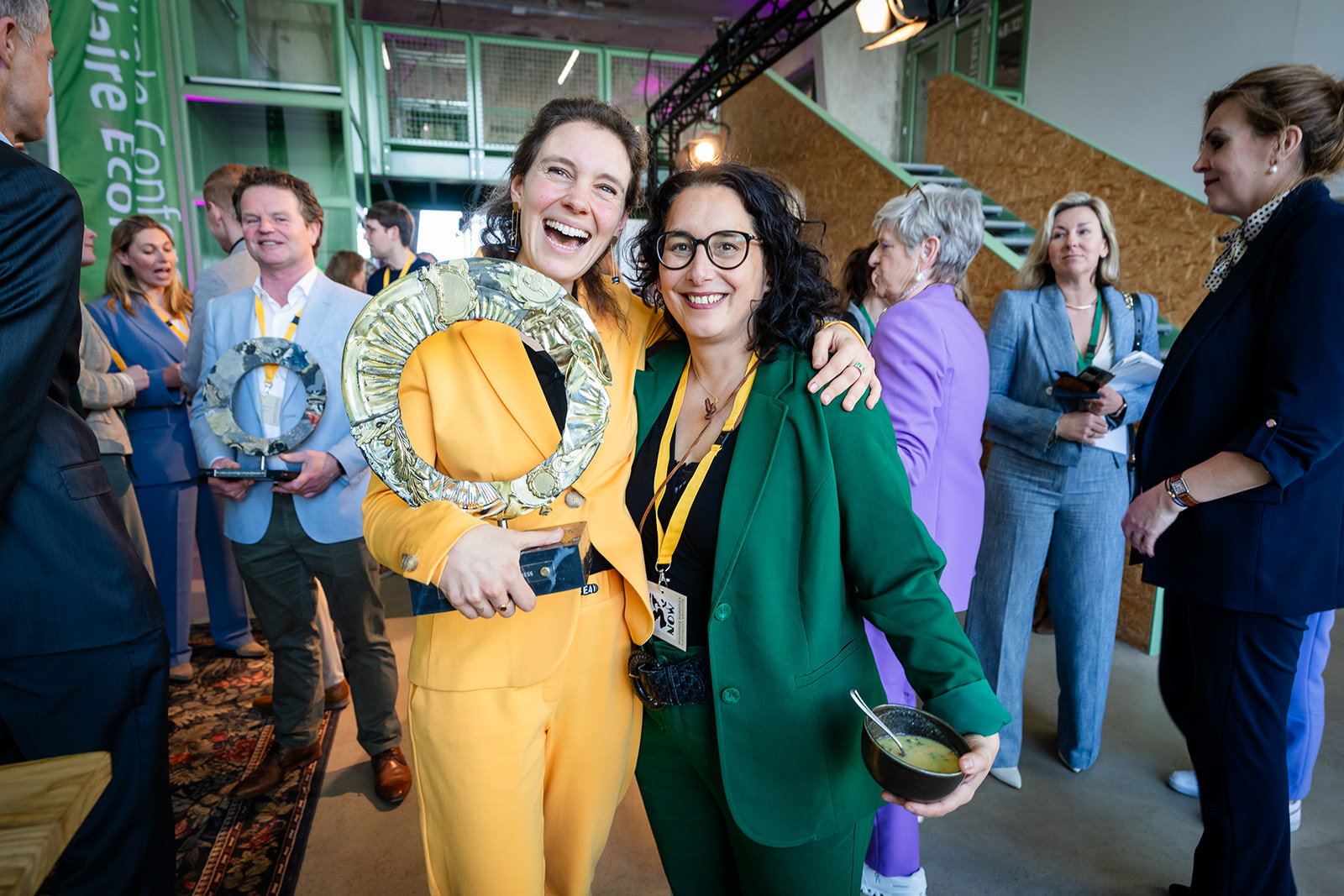 twee vrouwen in geel en groen pak lachen voor de camera met prijs